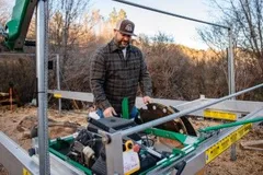 A man in a plaid jacket and hat operates a green and silver portable sawmill outdoors, surrounded by trees and dry grass.