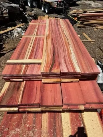 Long, wide wooden boards with striking red, pink, and cream stripes are stacked outdoors on the ground, surrounded by other pieces of wood and construction materials.